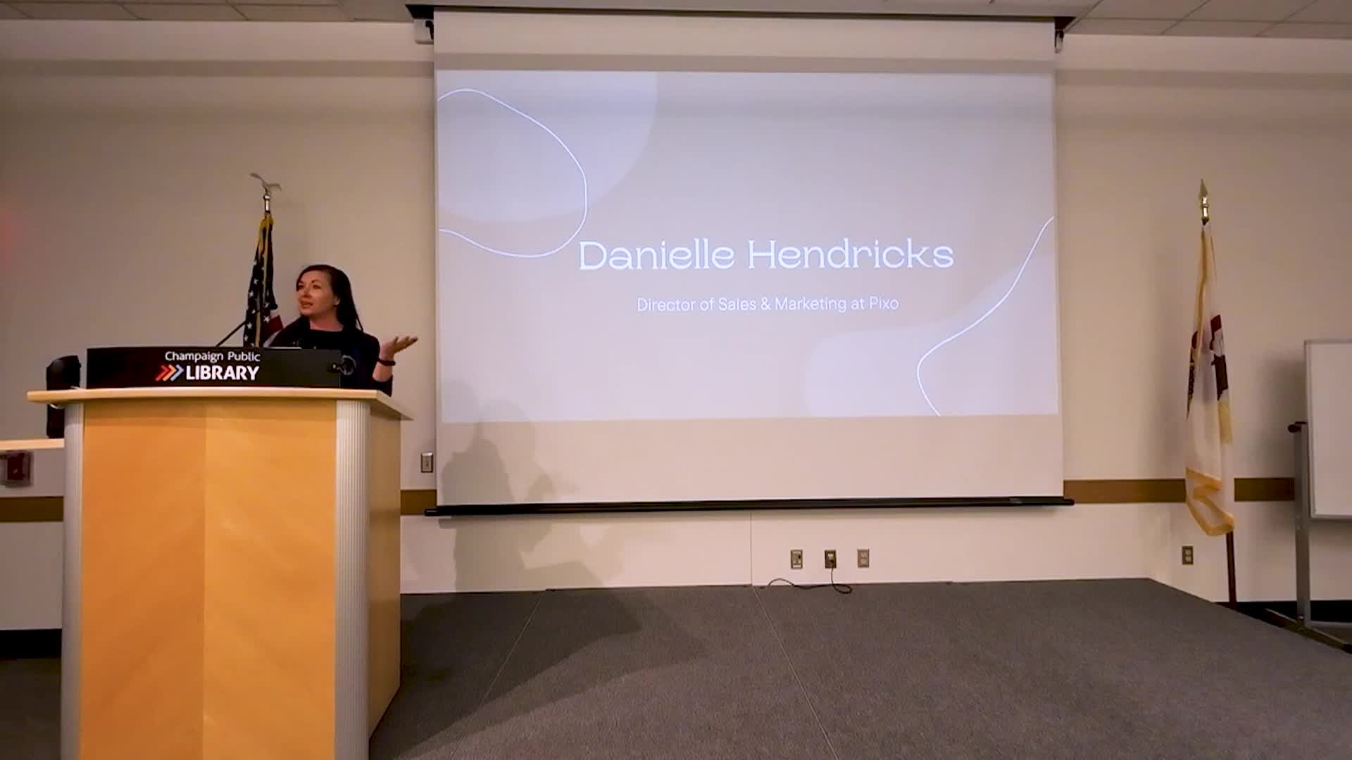 Wide shot of front of room with the speaker and a projector screen. 