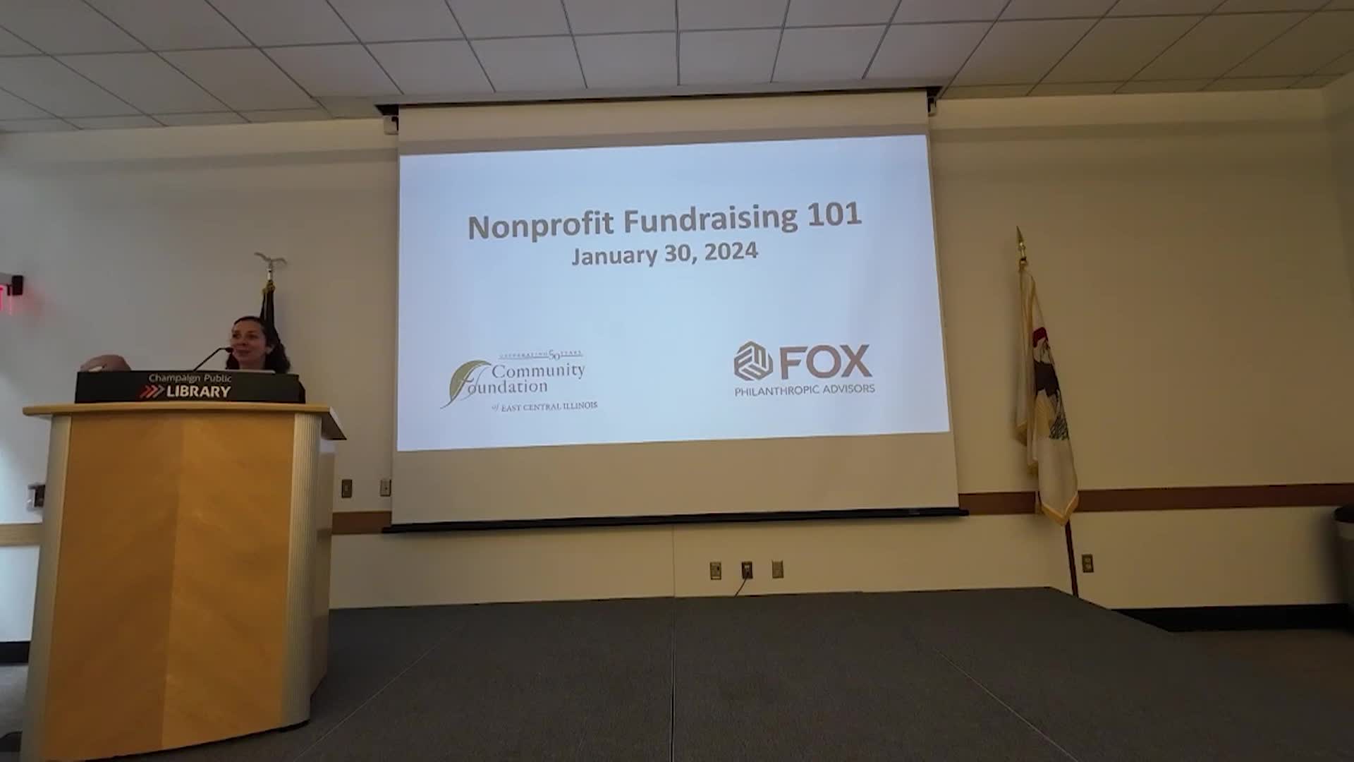 Wide shot of the front of the room with the speaker standing at a podium and a projector screen next to them. 